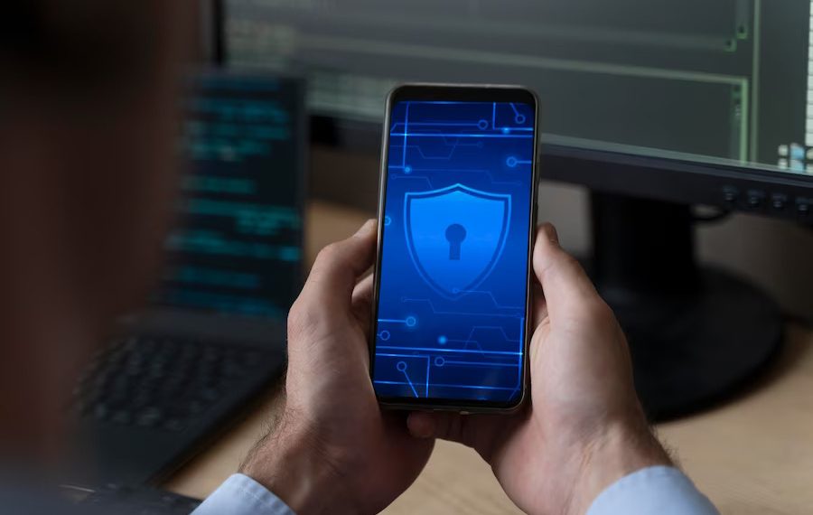  A person is holding a smartphone with a shield icon on the screen, which represents the search query "Aplikasi chat anonim terbaik untuk android".